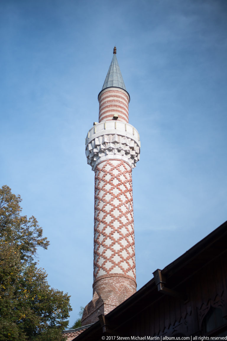 Patterned red-and-white brick minaret with projecting balcony and conical cap rising above a roof in Plovdiv.