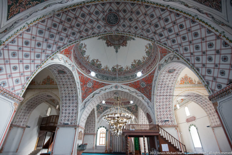 Decorated mosque interior with patterned arches, floral painting, chandelier, tall windows, and a wooden screened stair structure.