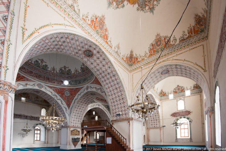Oblique mosque interior with painted intersecting arches, floral ornament, chandelier, windows, and dark wooden stair enclosure.