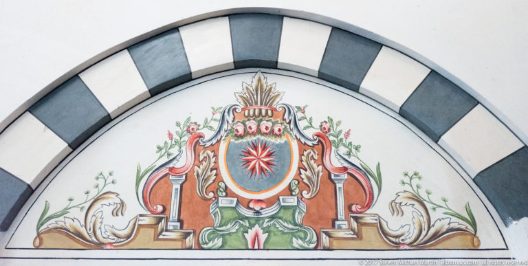 Colorful painted emblem with red shield, starburst, flowers, and scrolling forms inside a black-and-white arched border.