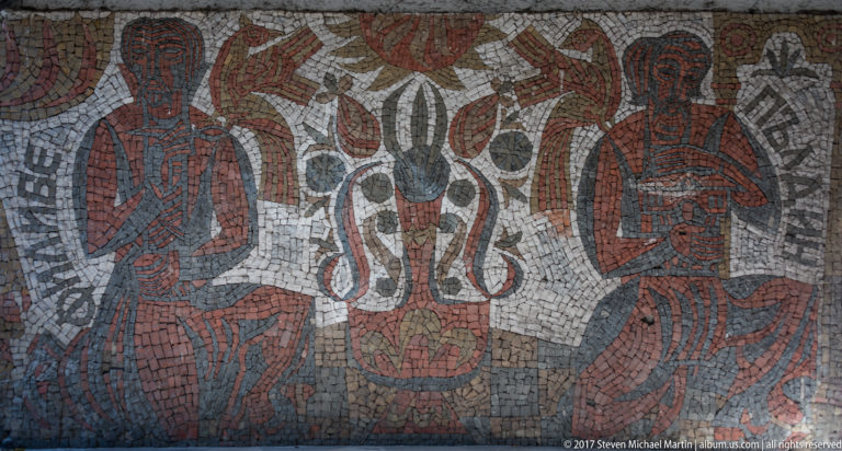 Mosaic mural with two seated red-brown figures flanking a symmetrical black-and-white animal-like motif.