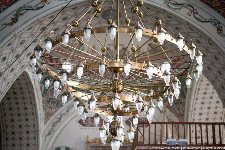 Large multi-tier brass chandelier with many white glass lamps hanging before patterned mosque arches.