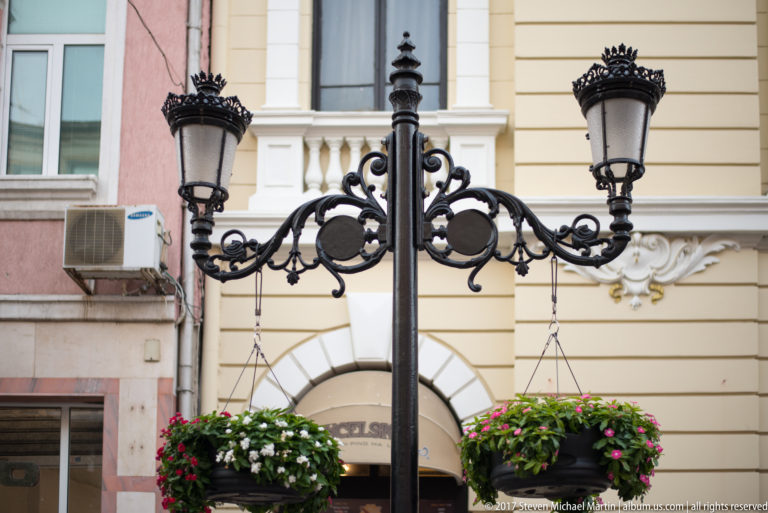Ornate black double streetlamp with scrolling ironwork and hanging flower baskets against a cream historic façade.