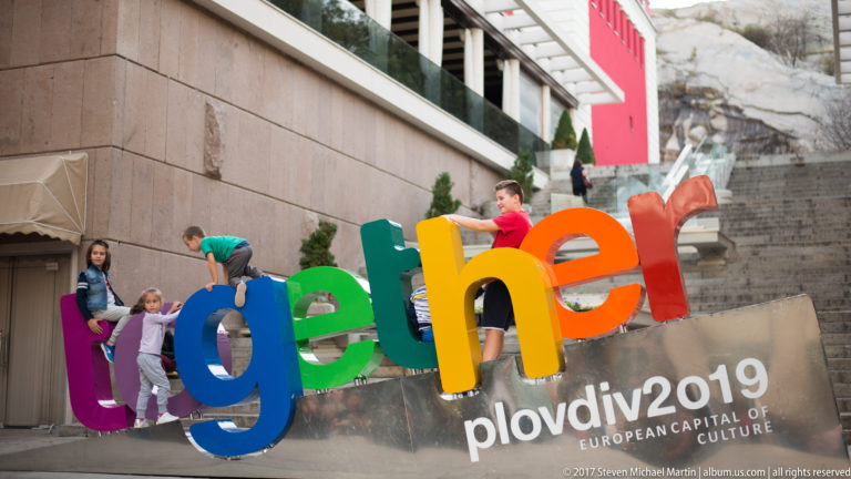 Children playing around giant rainbow “together” letters with Plovdiv 2019 European Capital of Culture text below.