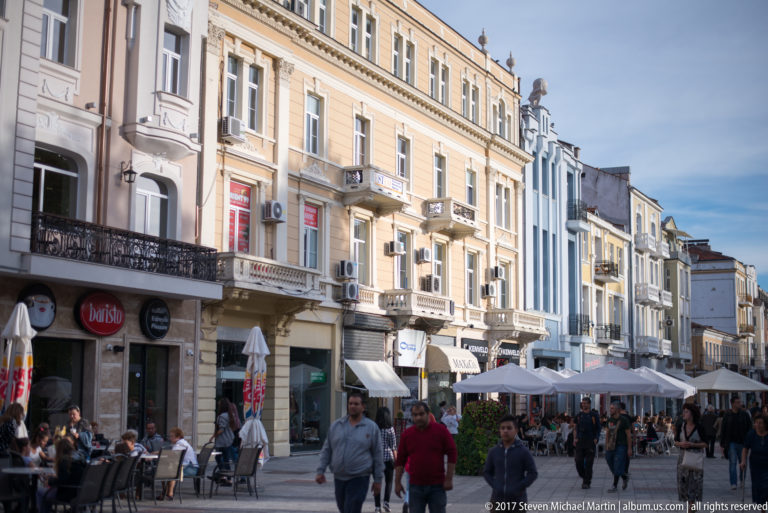 Pedestrians and café tables along a central Plovdiv street lined with multi-story historic façades and balconies.