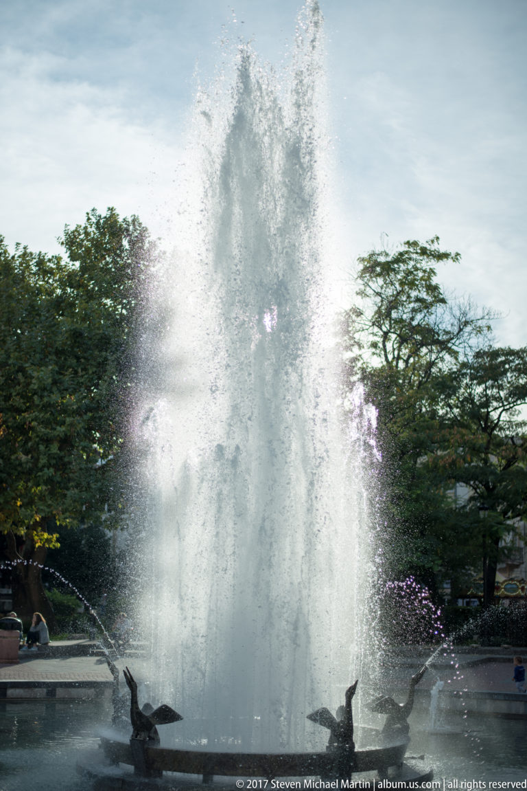 Tall white fountain jets and lower sprays rising from a basin against dense green trees in a Plovdiv park.