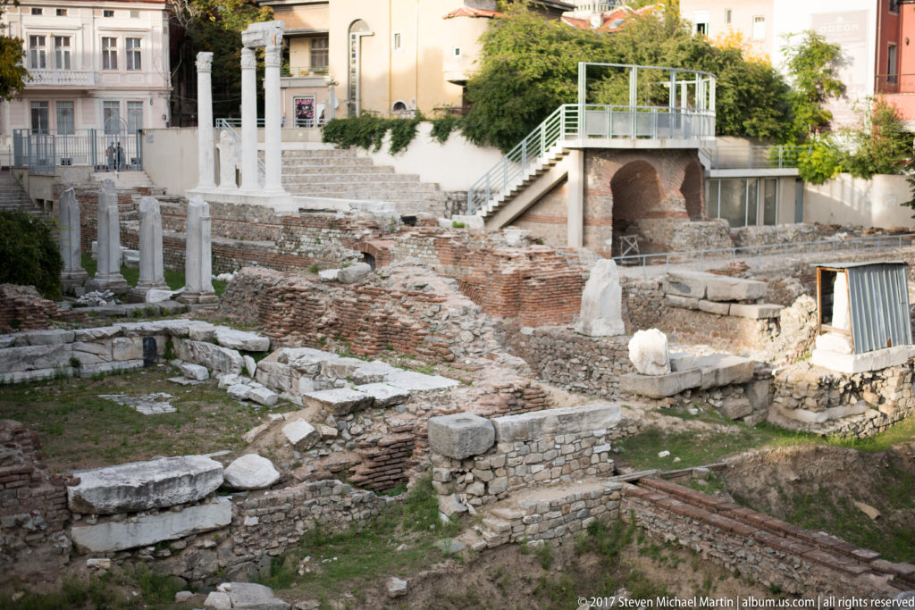 Excavated brick and stone walls with column fragments below modern buildings, stairs, and railings in central Plovdiv.