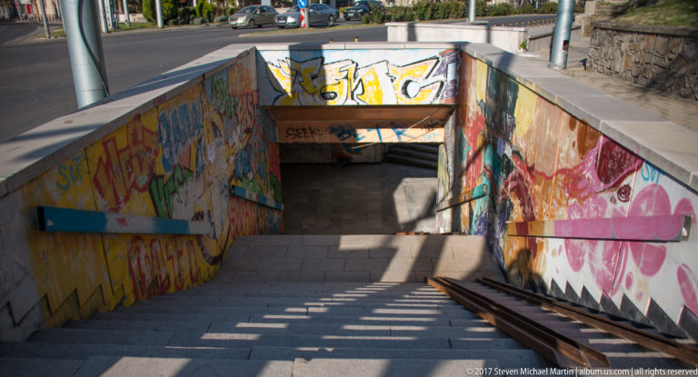 Outdoor stairs descending into a graffiti-covered pedestrian underpass, with handrails and strong diagonal shadows.