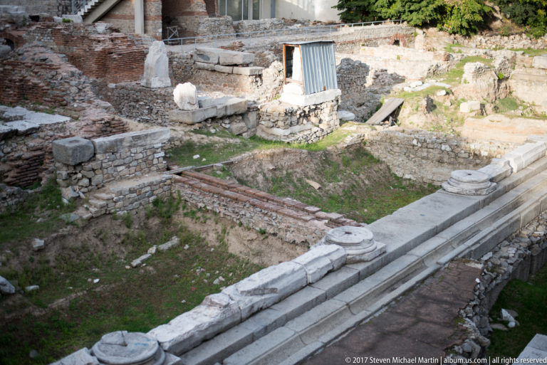 Pale stone steps bordering excavated brick-and-stone foundations with scattered architectural fragments and grass.