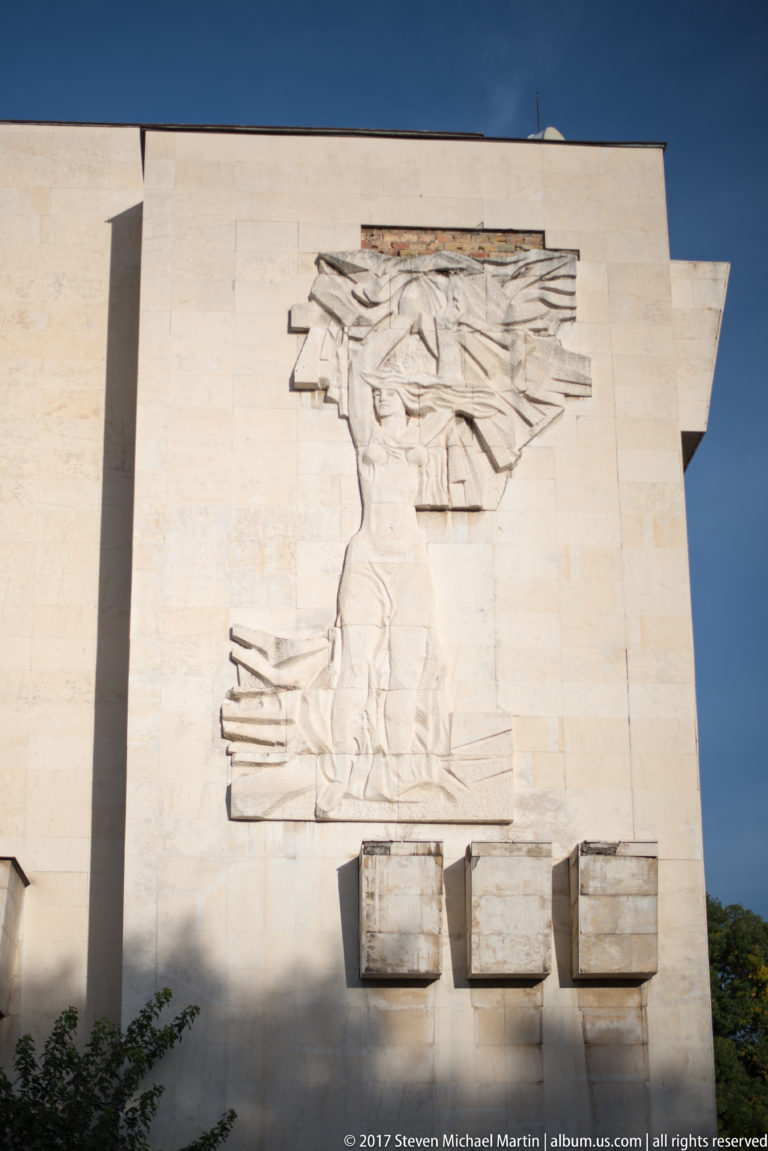 Large pale abstract figurative relief on a blank building wall, with three rectangular projecting blocks beneath.