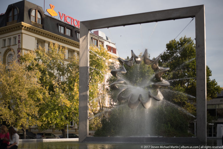 Spiked circular fountain sculpture suspended in a rectangular frame, spraying water into a pool amid trees.