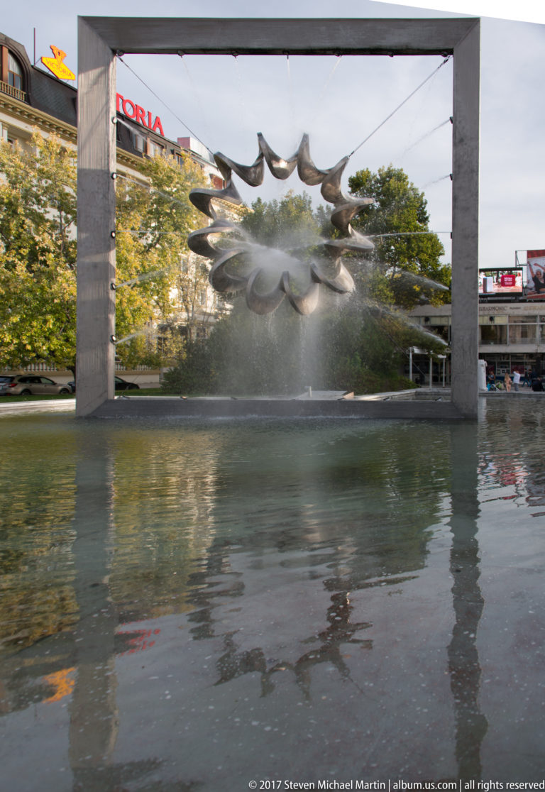 Wide view across a reflecting pool toward a framed spiked fountain sculpture spraying a fine curtain of water.