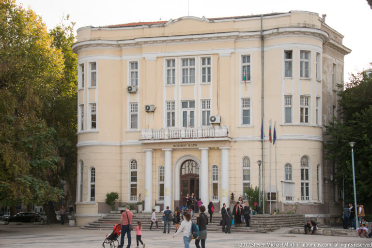 Pale academy building with tall windows, central arched entrance, balcony, flags, and people gathered on the steps.