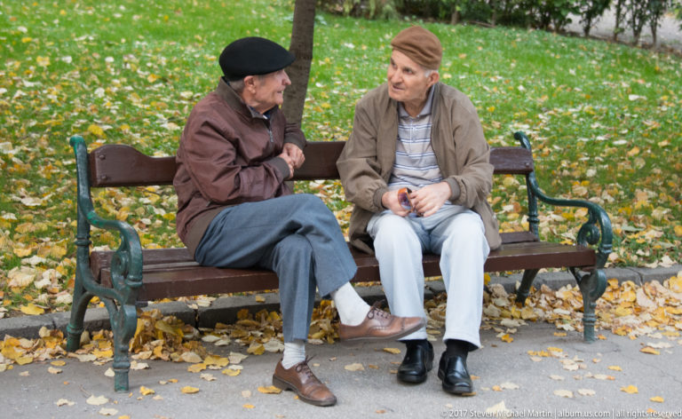 Two older men in caps talking on a wooden park bench surrounded by fallen autumn leaves.