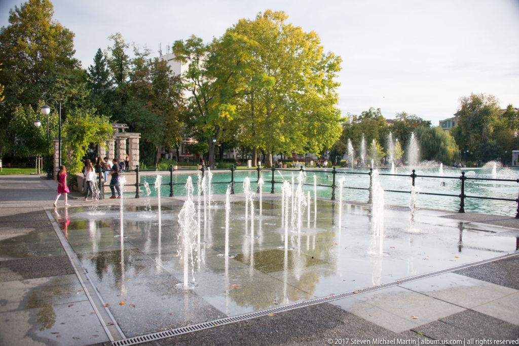 Rows of ground-level fountain jets on wet paving beside a larger lake fountain, with visitors and trees beyond.