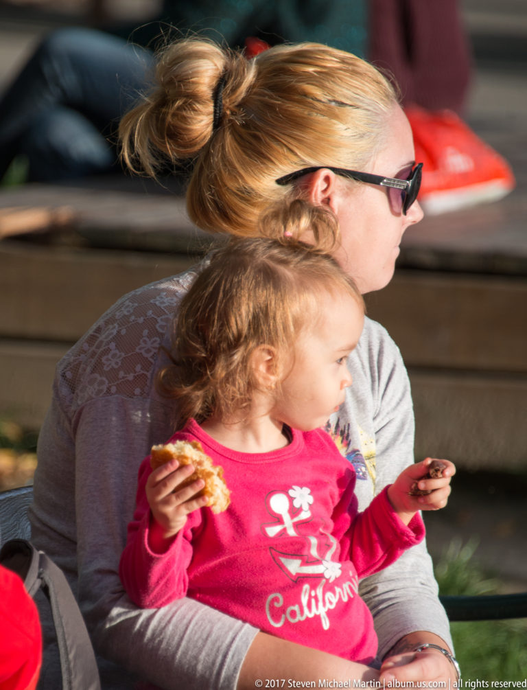 Woman in sunglasses holding a young child in a red shirt who is eating while seated outdoors.