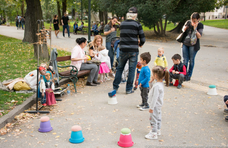 Children and adults gathered on a park path around colorful plastic markers and small handmade figures near a bench.