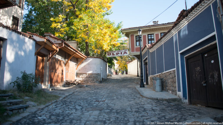 Steep cobblestone lane in Old Plovdiv between traditional houses, with trees and a sign spanning the street.