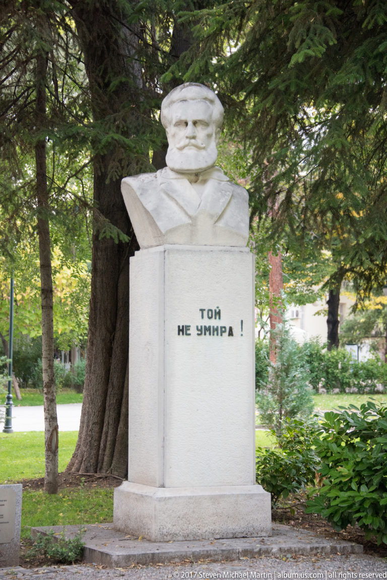 Stone bust of a mustached man on a pedestal inscribed in Bulgarian “ТОЙ НЕ УМИРА!” beneath trees.