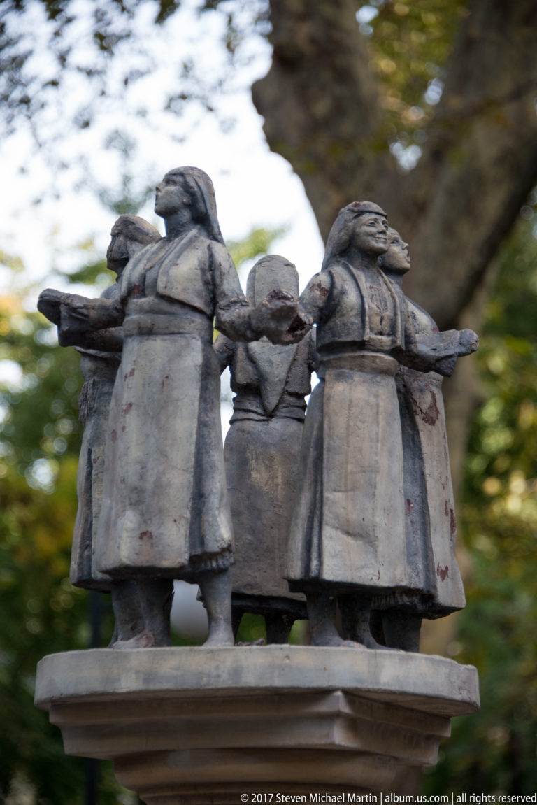 Bronze sculpture group of women in broad traditional-style dresses gathered around a central object with arms extended.