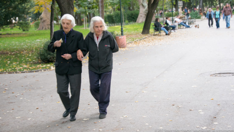 Two elderly women in dark clothing walking arm in arm along a park path with benches and trees behind.