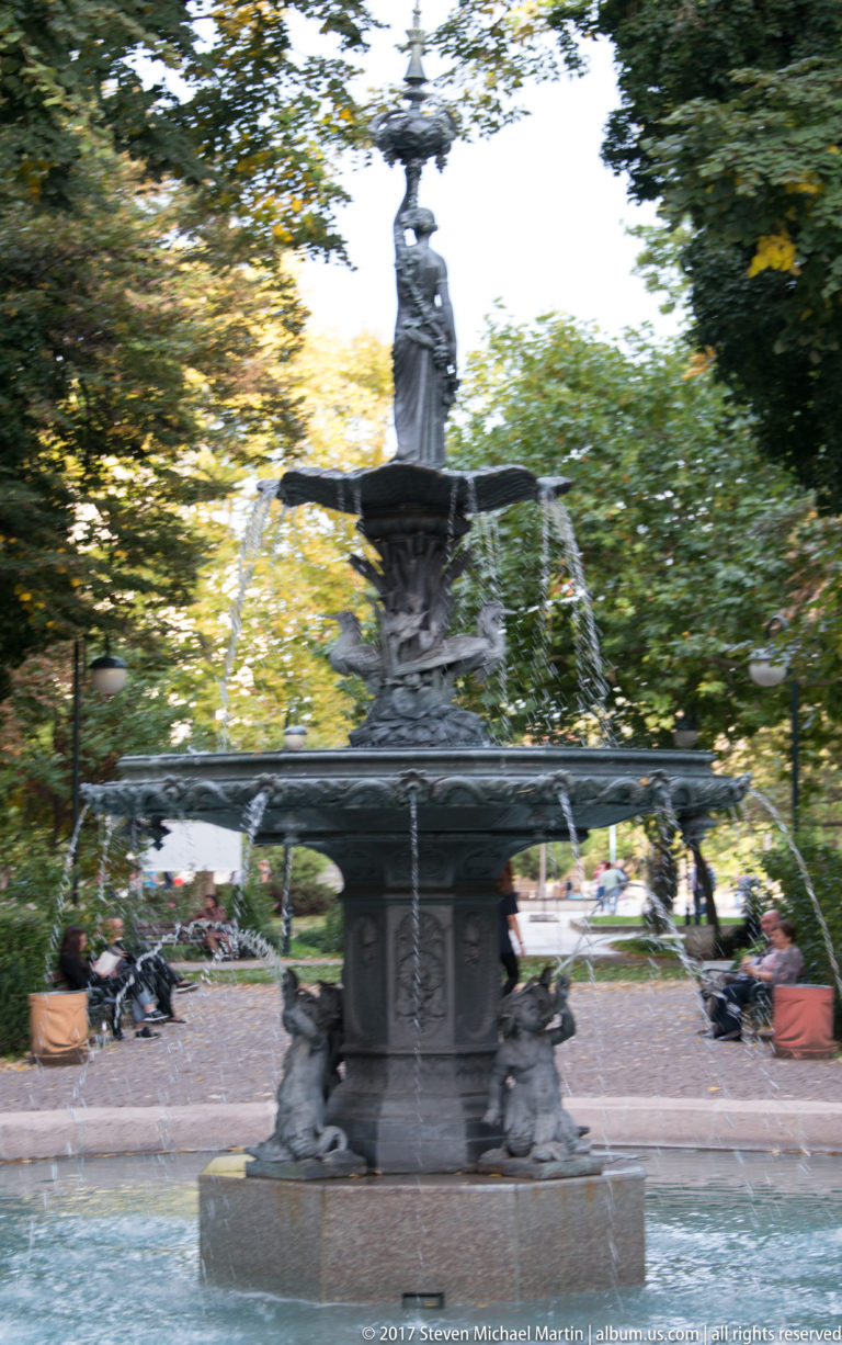 Dark tiered fountain with a standing female figure on top, arcing water jets, and sculpted figures around the base.