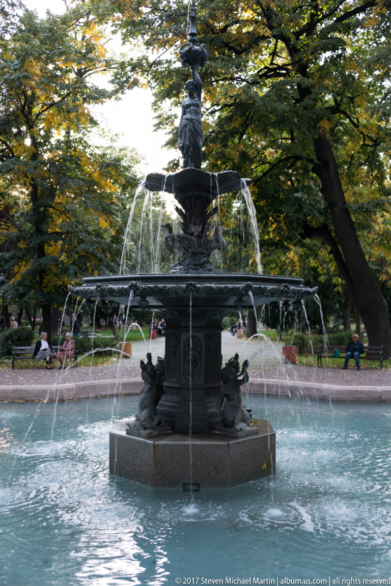 Full frontal view of an ornate dark fountain in a turquoise pool, with tiered water jets and a female figure above.