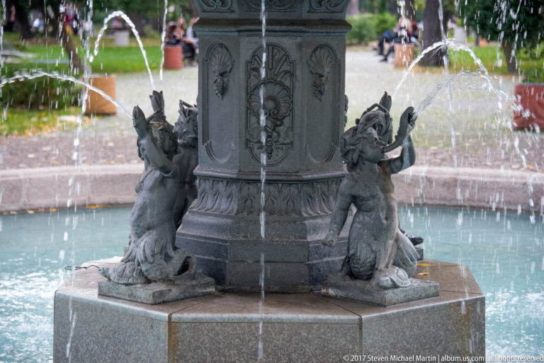 Close view of dark kneeling sculpted figures around a fountain pedestal with water falling behind them.