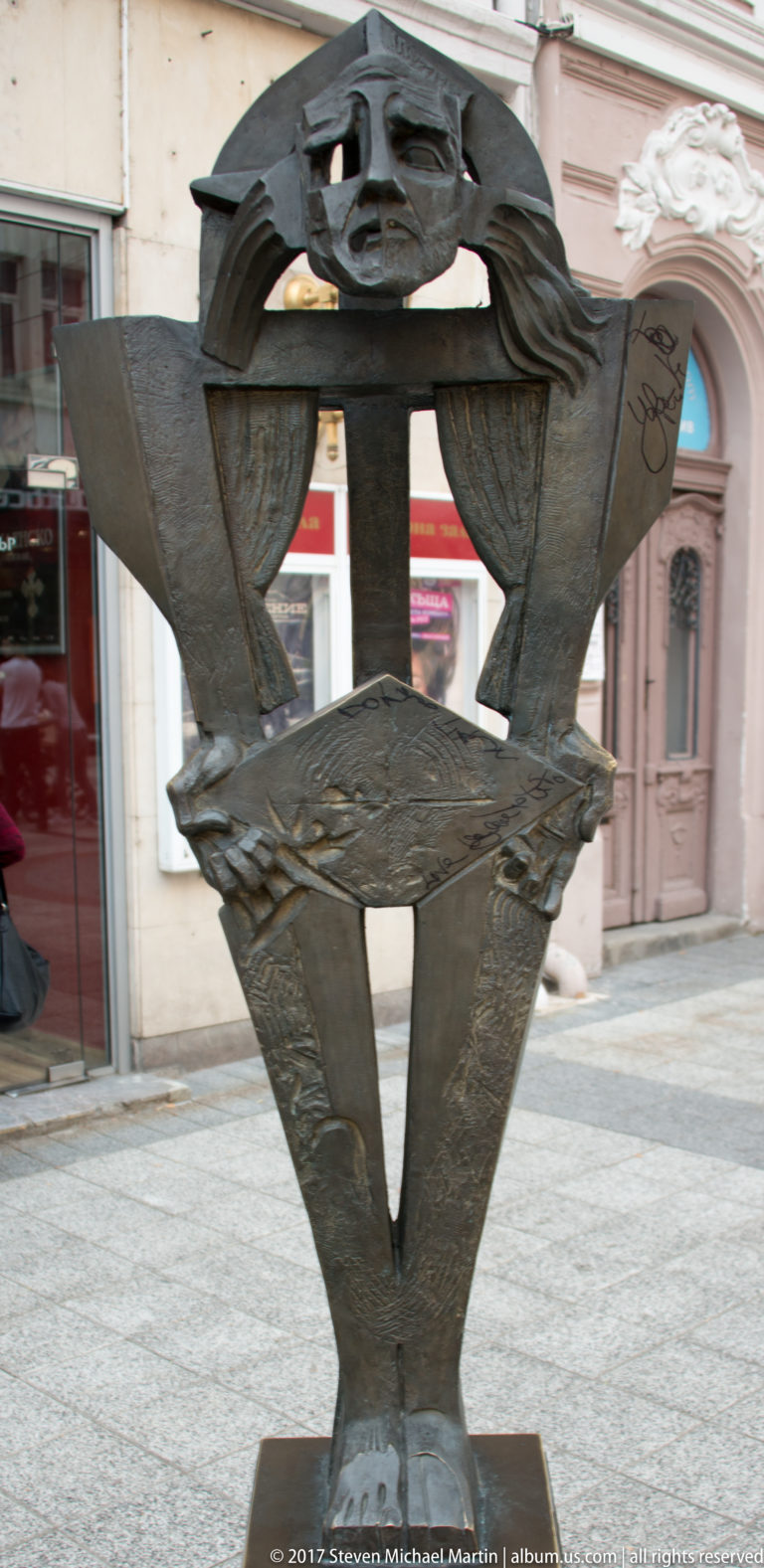 Dark abstract metal sculpture with triangular planes and curved openings on a pedestal along a Plovdiv pedestrian street.