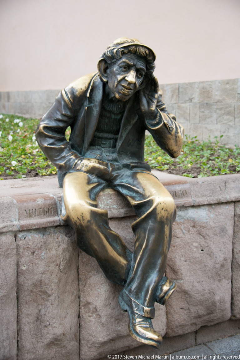 Life-size bronze seated man leaning his head on one hand while resting on a low stone ledge.