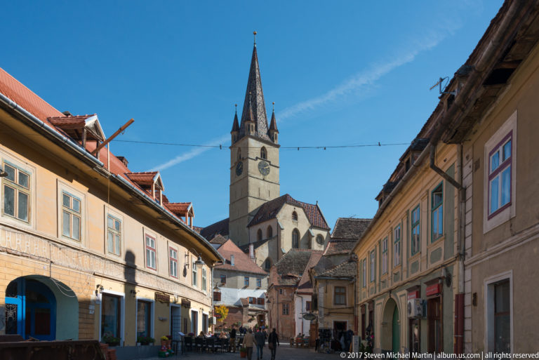 Historic street in Sibiu leading toward a tall church tower with pointed spire, red roofs, and pedestrians.