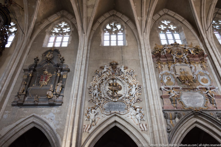 Three ornate wall epitaphs with coats of arms and inscriptions between Gothic arches and tall traceried windows.