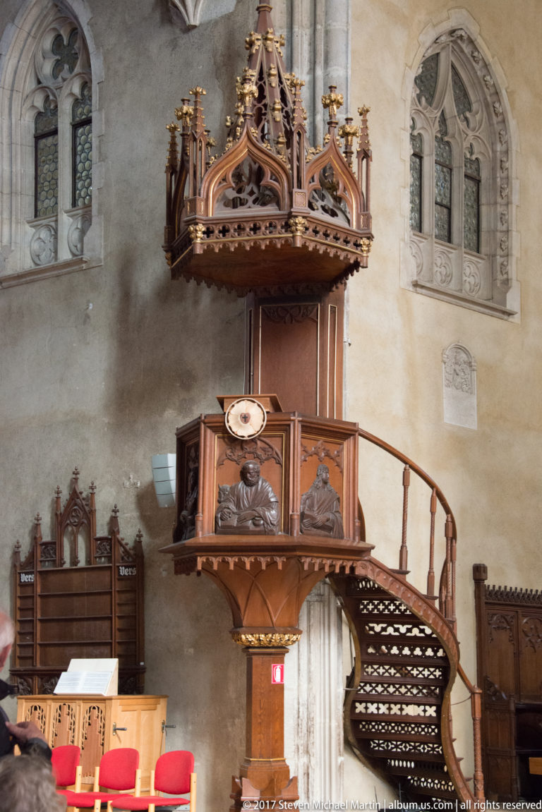 Carved wooden church pulpit with curved staircase, panelled base, and ornate Gothic-style canopy.