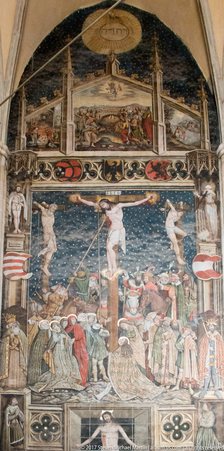 Weathered church mural with a central Crucifixion scene, figures, heraldic bands, and multiple painted registers.