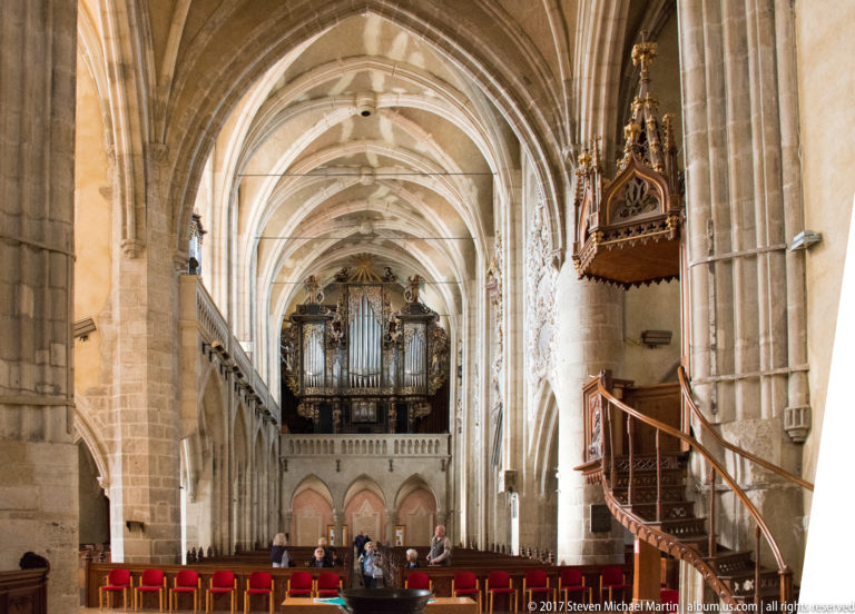 Long Gothic church nave with ribbed vaults, stone piers, pipe organ, carved pulpit, and rows of red seats.