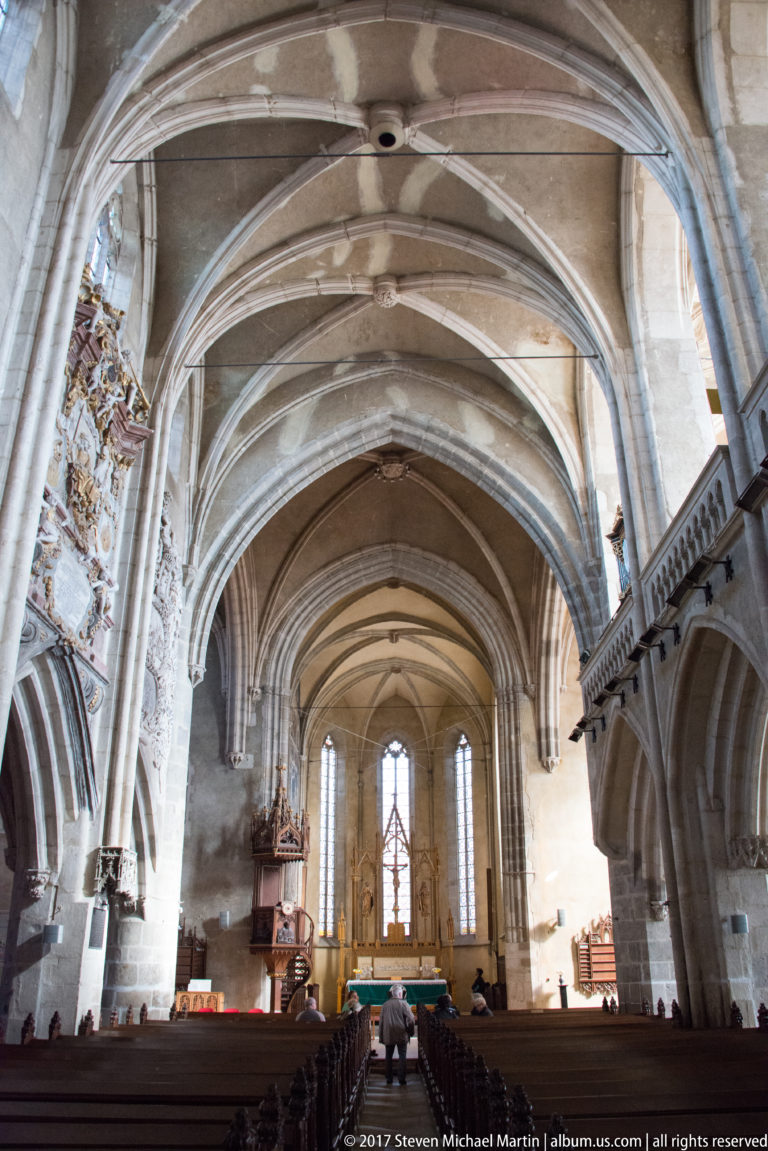 Gothic church nave with ribbed vaults, arcades, tall sanctuary windows, altar, and a few visitors.