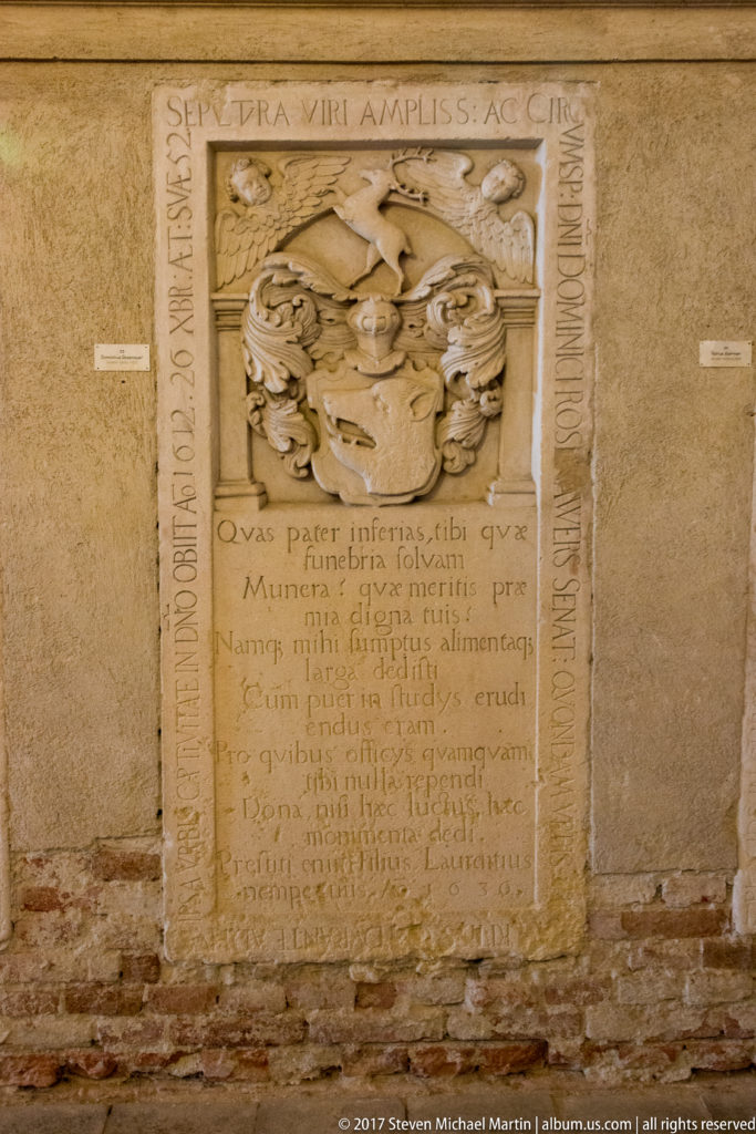 Upright stone funerary slab with carved coat of arms, long Latin inscription, border, and worn surface.