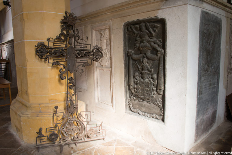 Wrought-iron cross beside carved stone memorial slabs in the corner of a historic church interior.