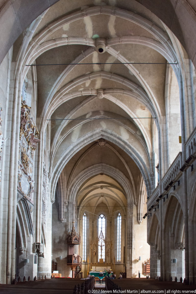 Wide Gothic church nave looking toward the sanctuary, with ribbed vaults, stone piers, side arcades, and pews.