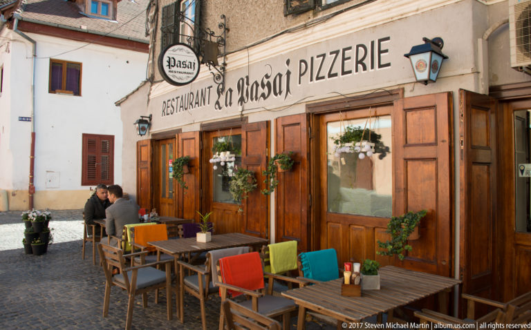 Pasaj restaurant frontage with wooden doors, hanging flowers, outdoor tables, and colorful chair cushions on a cobbled lane.