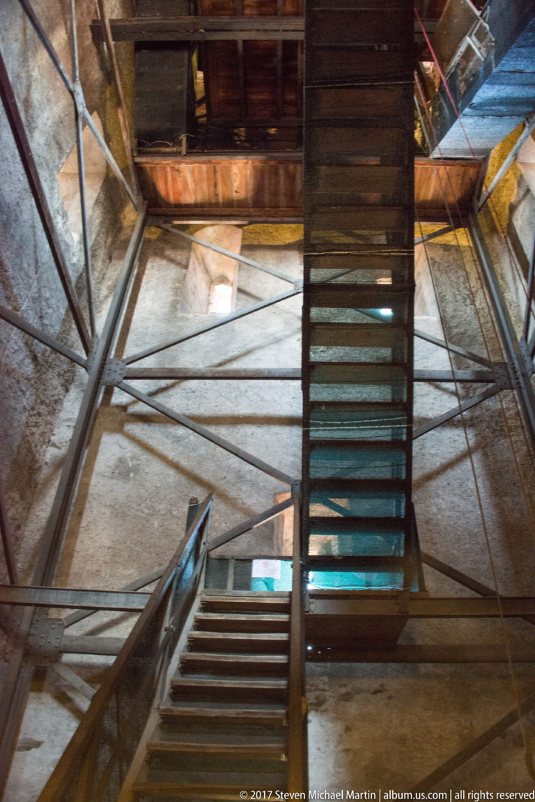 Interior of a historic tower with steep wooden stairs, landings, structural braces, and daylight entering through openings.