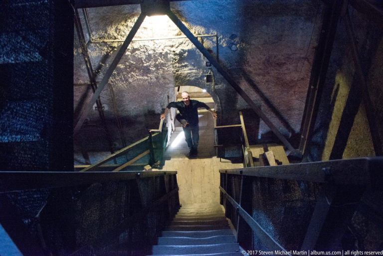 Visitor climbing steep stairs inside a dark historic tower with timber framing and a bright opening behind.