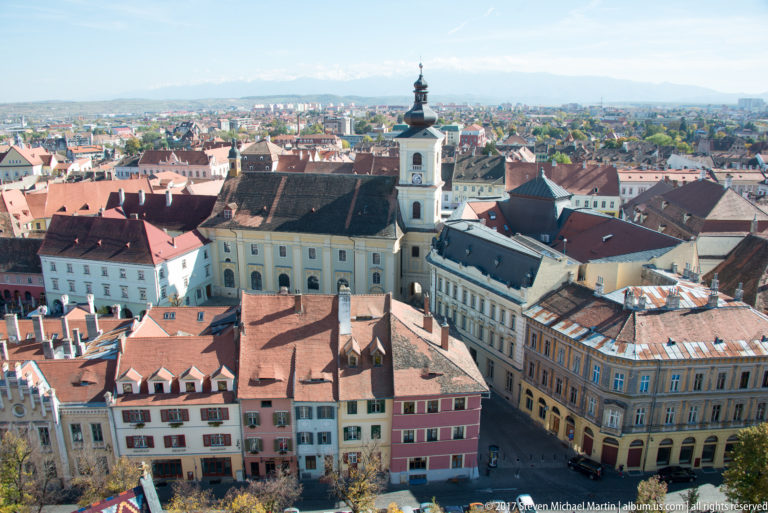 Elevated Sibiu cityscape with red-tiled roofs, pastel façades, a white church and tower, and distant hills.