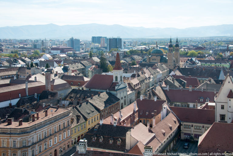 Elevated panorama over Sibiu’s historic roofs and church towers toward the modern city and distant mountain ridge.