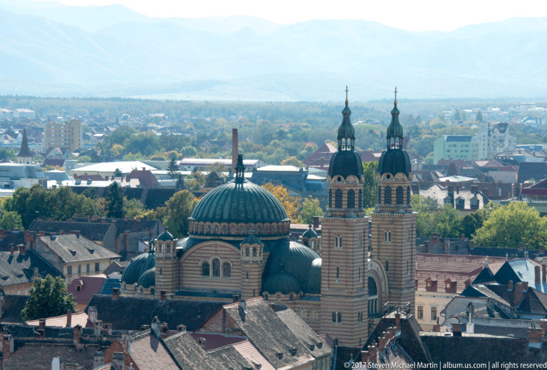 Large domed cathedral in Sibiu with twin bell towers, striped masonry, smaller domes, and city rooftops around it.
