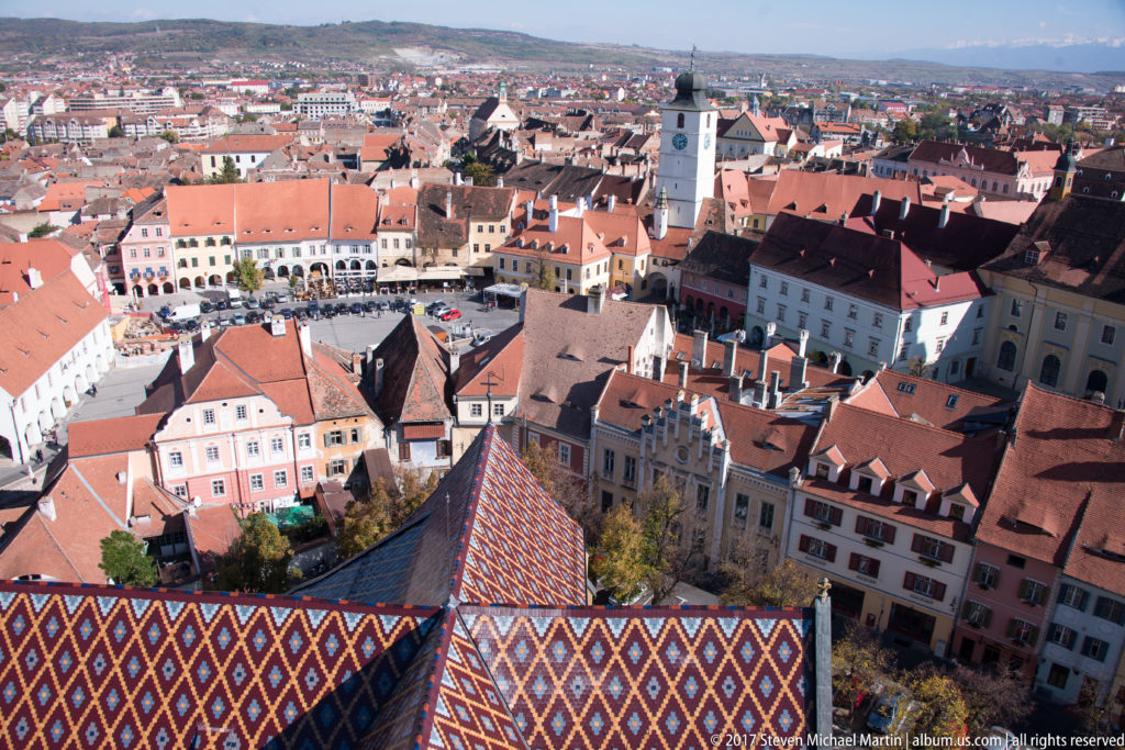 High view over a colorful patterned roof toward Sibiu’s historic square, church towers, and dense red-roofed old town.