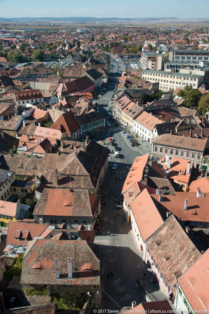 Elevated view over dense Sibiu streets, courtyards, and red-brown tiled roofs with larger modern buildings beyond.