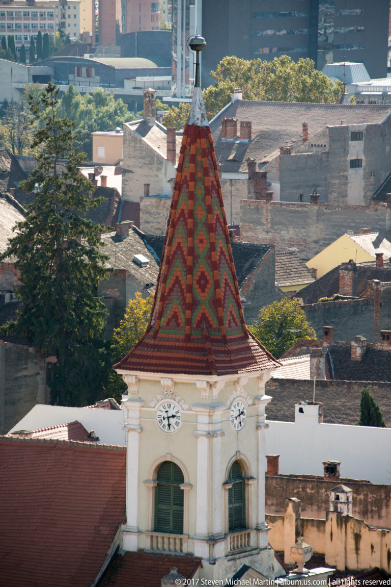 Tower with colorful geometric tiled roof, paired clock faces, pale walls, and a slender finial above Sibiu rooftops.