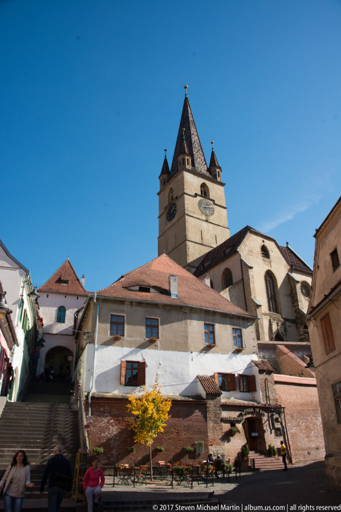 Square church tower with clock faces and corner turrets above red roofs, steps, and old-town buildings in Sibiu.