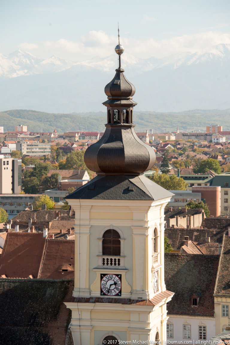 White bell tower with clock, arched opening, dark onion-shaped roof, and mountains beyond Sibiu.